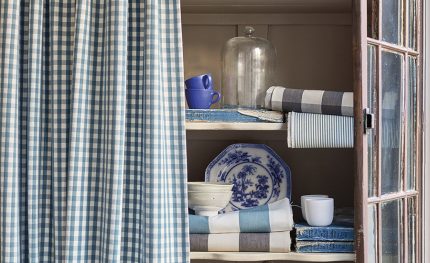 Tende e cuscini nel tessuto a quadretti Elmer di Romo, collezione Kemble, accanto a una vetrina con ceramiche. In vendita c’è soltanto il tessuto al metro: arredi e complementi della scena non sono compresi.